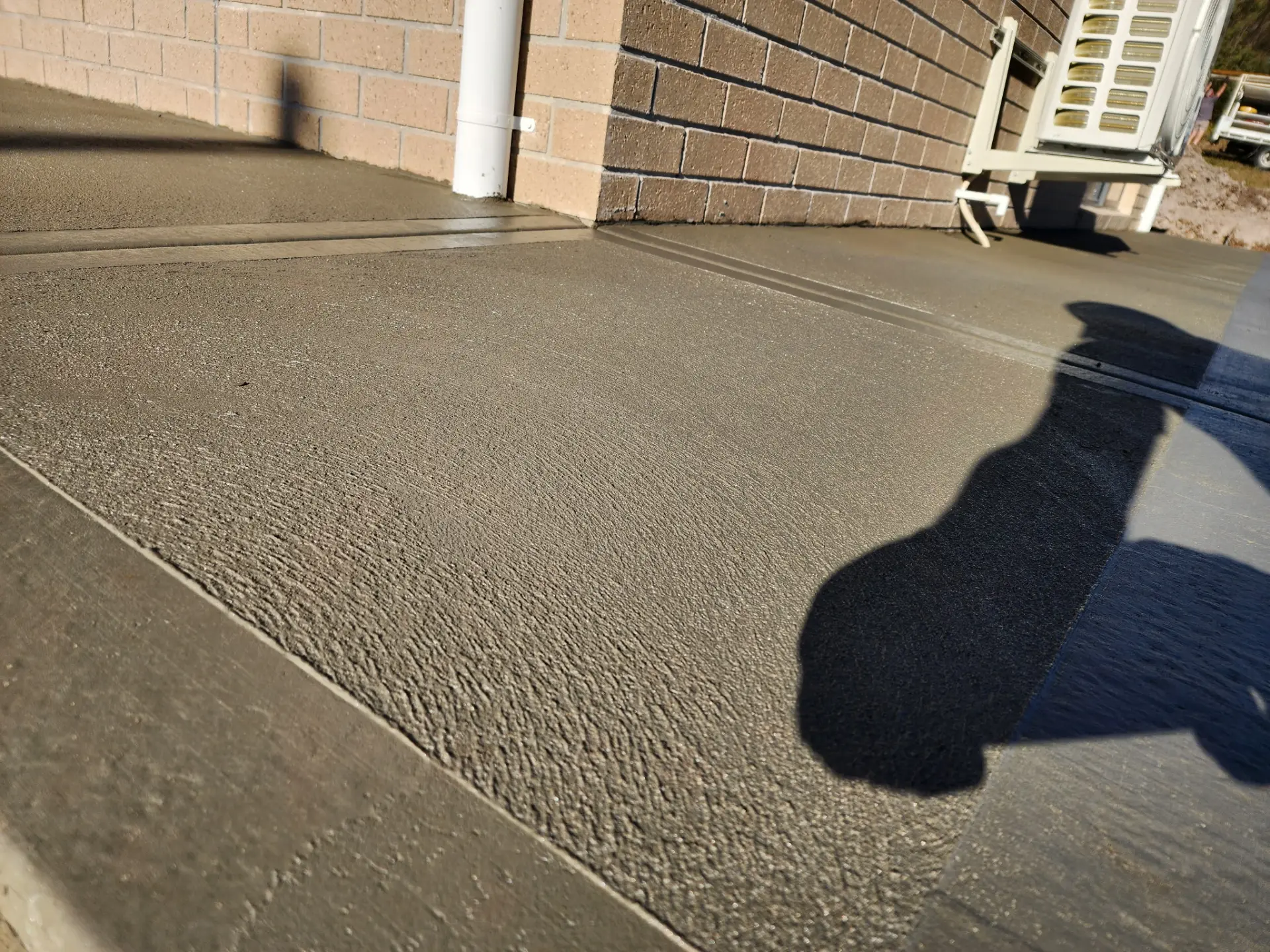 A freshly floated concrete slab beside a brick wall with the concreter's shadow across the surface