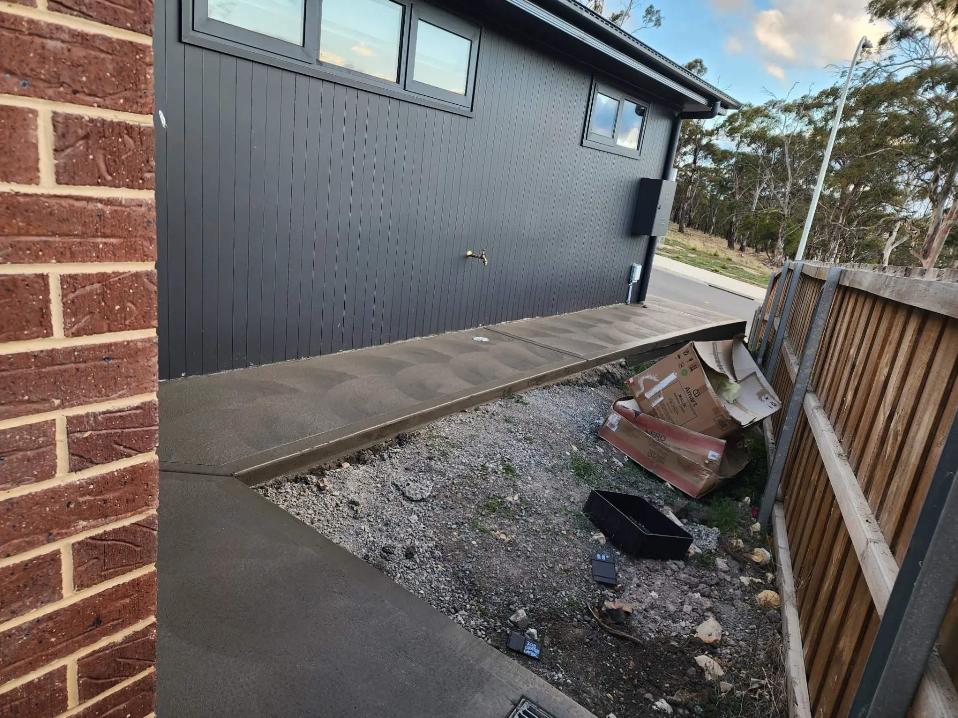 A concrete path and outdoor area laid beside a dark-clad house and paling fence