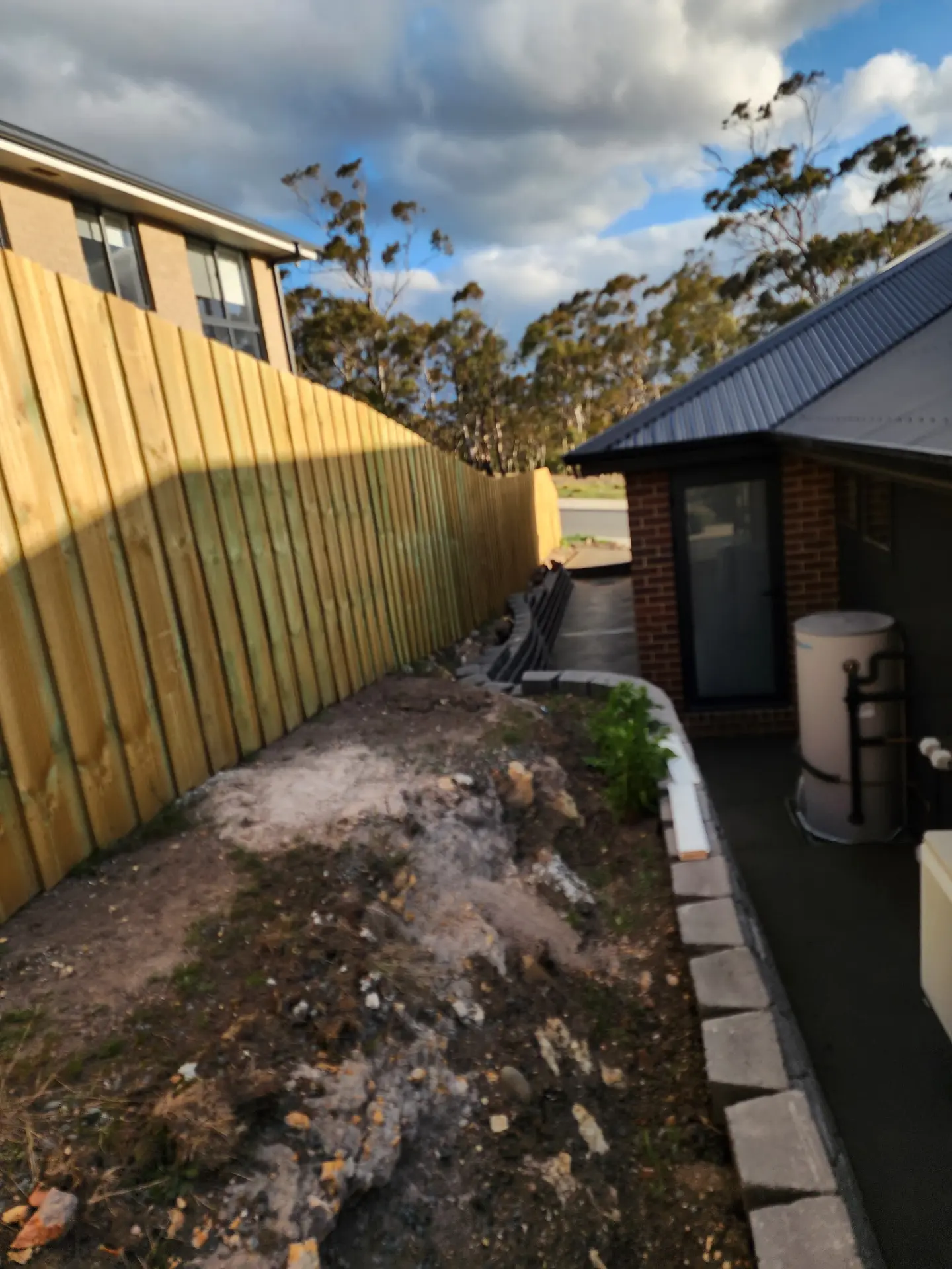 A finished side path running between a timber fence and a brick house on a sloping block