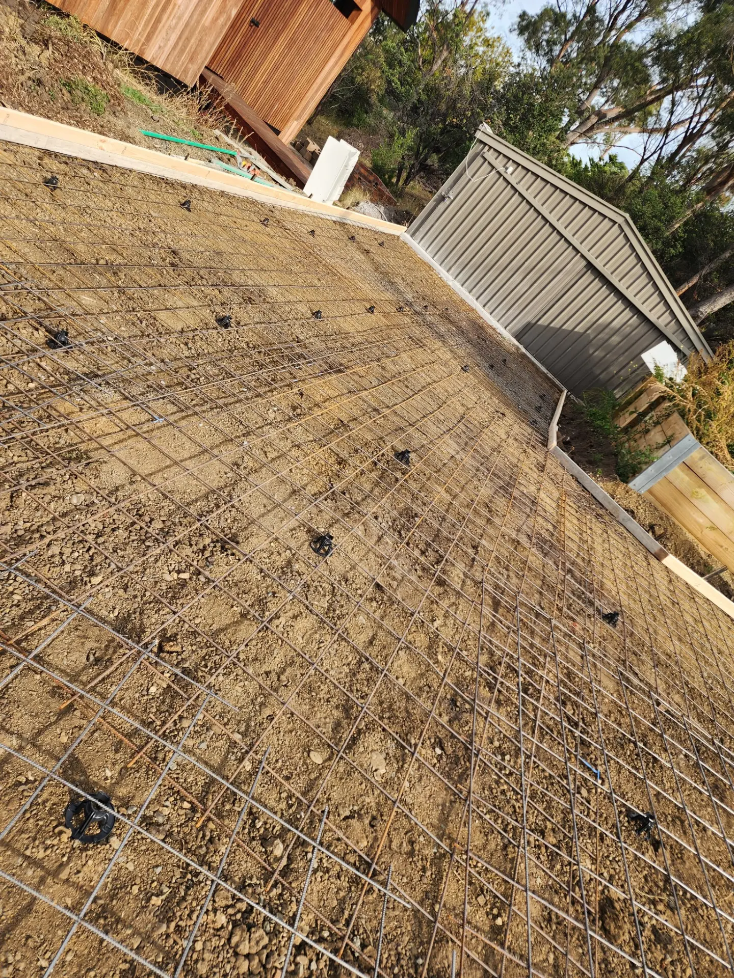 Reinforcing mesh sitting on bar chairs across a prepared pad beside a shed