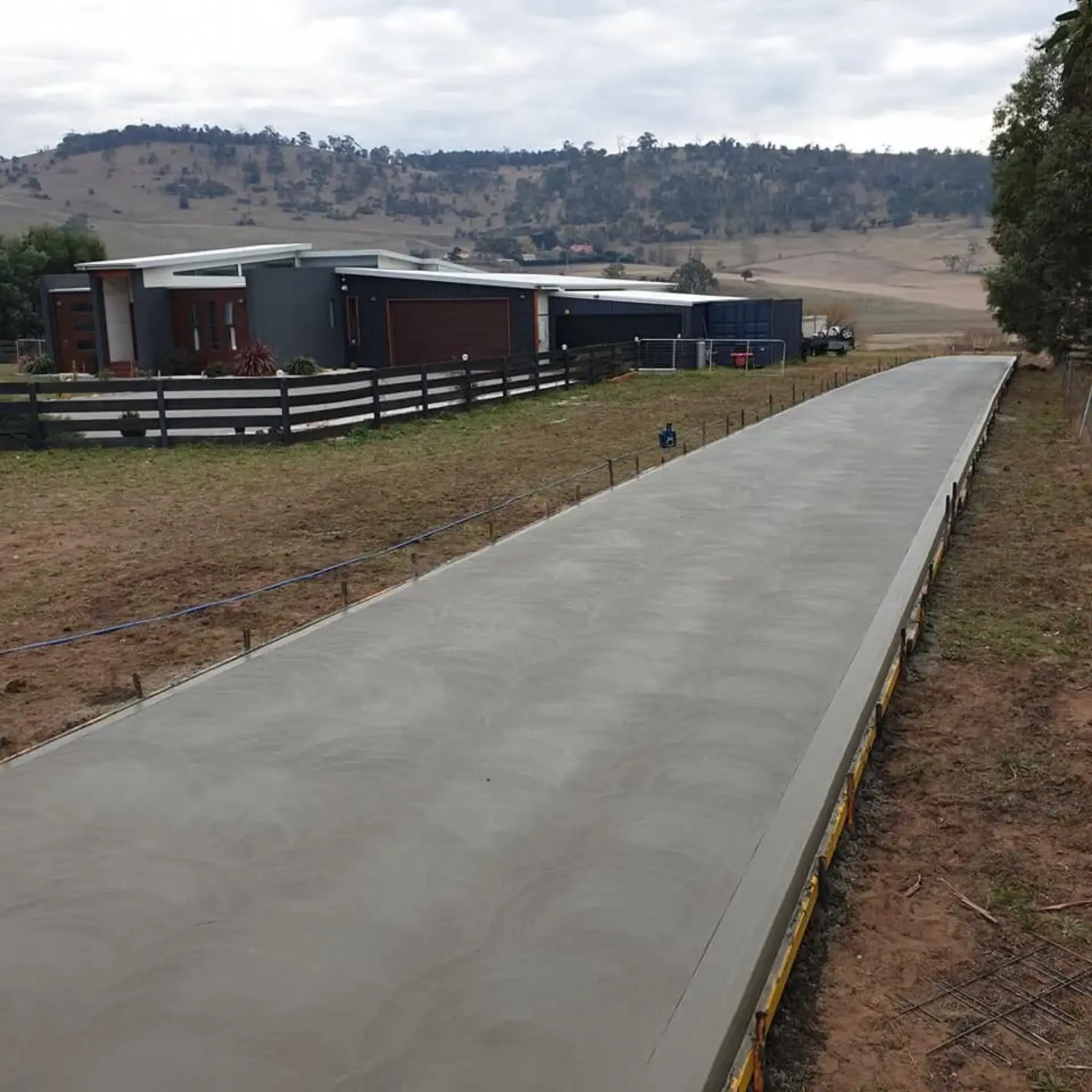 A long finished concrete driveway running beside a paling fence