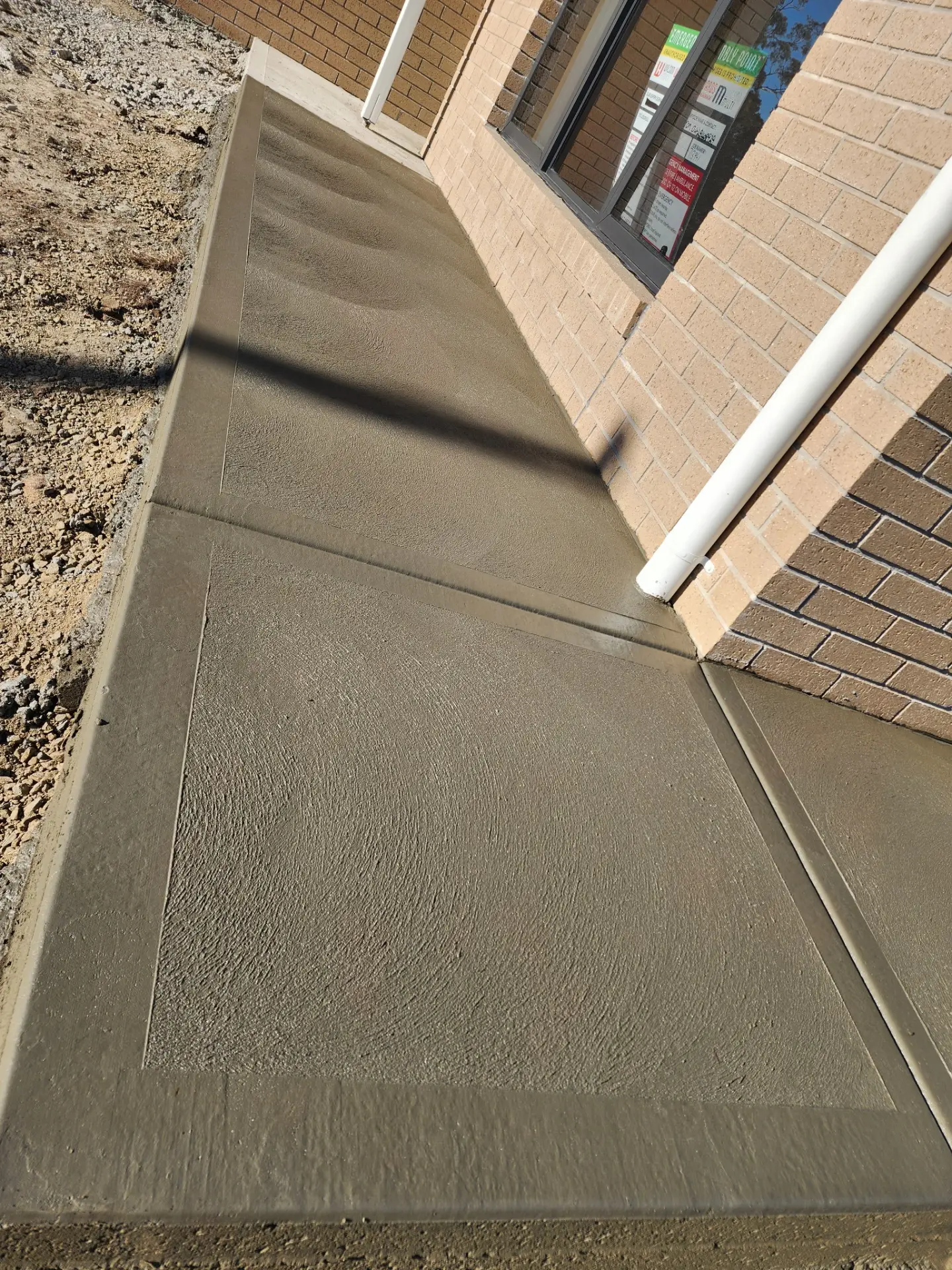 A fresh concrete path poured around the corner of a brick house beside a gravel strip