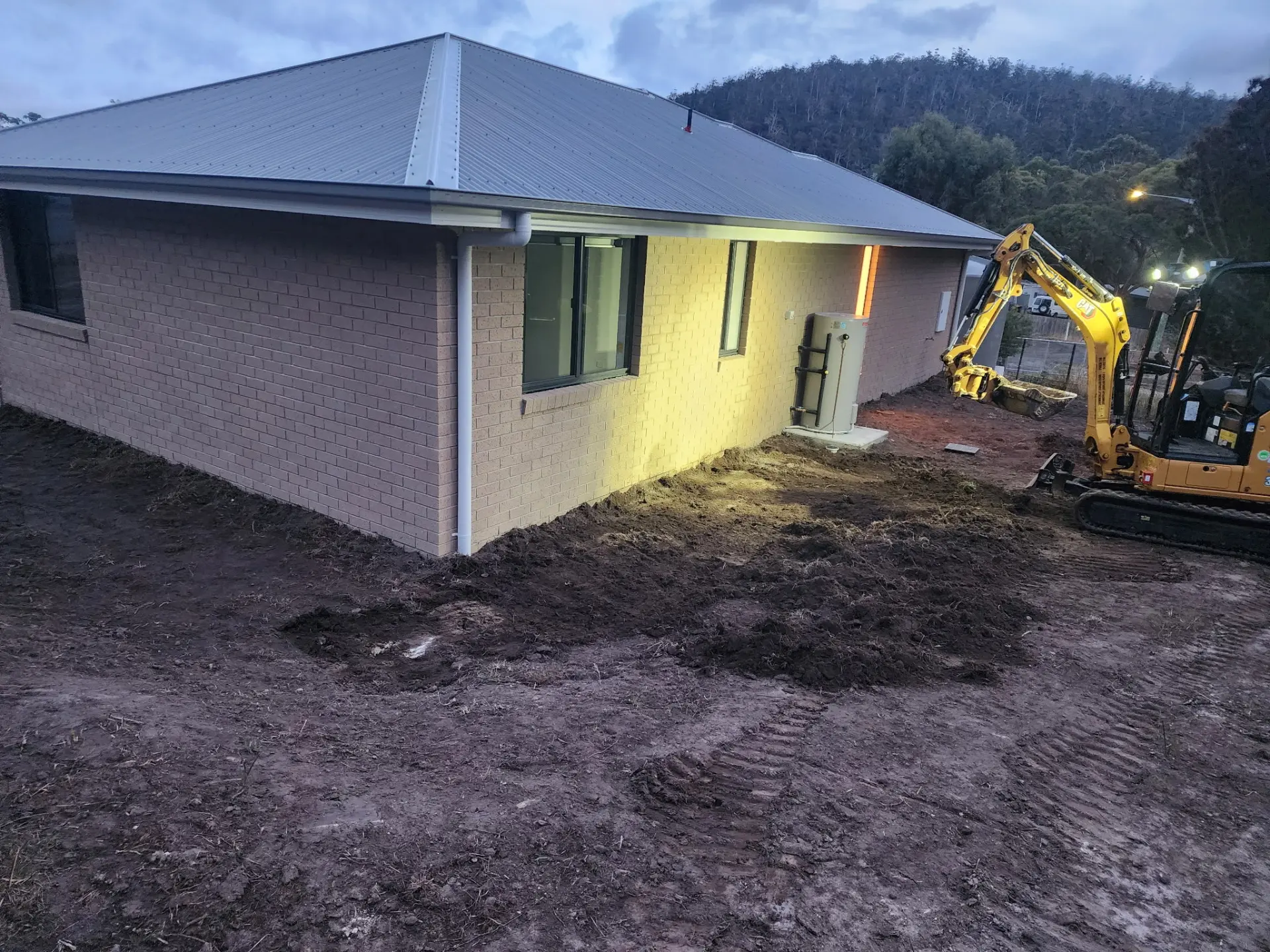 An excavator working ground beside a rendered house on a semi-rural block at dusk