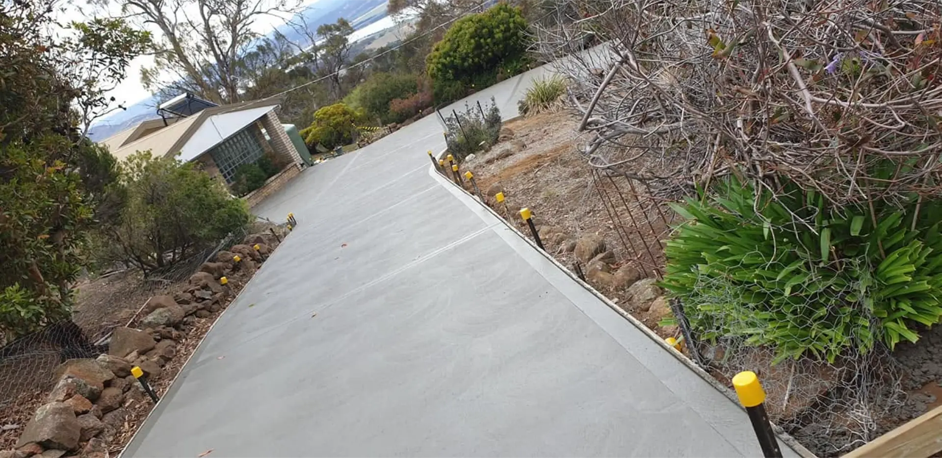 A newly poured plain concrete driveway sloping up past bollard markers to a home